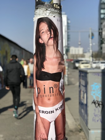 A poster of a young woman wearing a black top and white underwear with the text 'HEROIN KIDS' is wrapped around a pole on a city street. In the background, there are blurred images of people walking and urban infrastructure, including buildings and vehicles. Graffiti is visible on an electrical box nearby.