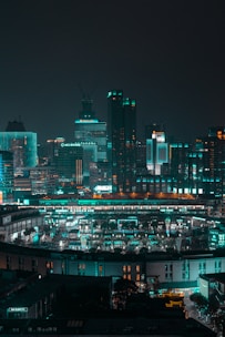 A nighttime urban cityscape with tall buildings and a vibrant, illuminated skyline. The buildings are adorned with teal and orange neon lights, creating a futuristic atmosphere. A densely packed city area with a circular formation is visible at the lower section, featuring well-lit streets and structures.