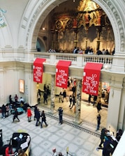 An ornate interior space with a high ceiling features intricate architectural details and a decorative, golden backdrop. Red banners with the text 'London Design Festival 2018' hang prominently. People are scattered throughout the area, walking and engaging in various activities.