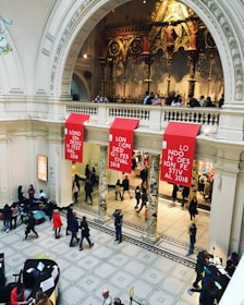 An ornate interior space with a high ceiling features intricate architectural details and a decorative, golden backdrop. Red banners with the text 'London Design Festival 2018' hang prominently. People are scattered throughout the area, walking and engaging in various activities.