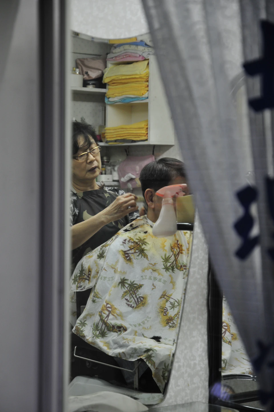 The image shows a woman cutting hair on another person's head, who is seated in front of her with their back to the camera. The woman holding the scissors appears to be focused and attentive while performing the haircut. The room has white walls and a window that allows natural light to enter, creating an atmosphere of comfort and relaxation for the client.