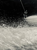 A person is wakeboarding on a water surface with a rope in hand. The image captures the spray and splash of water as the board cuts through the waves. The scene appears dynamic, emphasizing the motion and excitement of the activity. The background is dark, possibly trees or shadows, creating a contrast with the bright water in the foreground.