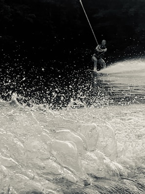 A person is wakeboarding on a water surface with a rope in hand. The image captures the spray and splash of water as the board cuts through the waves. The scene appears dynamic, emphasizing the motion and excitement of the activity. The background is dark, possibly trees or shadows, creating a contrast with the bright water in the foreground.