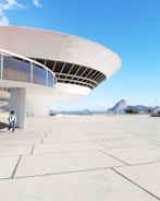 A futuristic, disc-shaped building with a sweeping overhang dominates the foreground. The structure features large windows and sleek, modern lines. In the background, a rocky outcrop and a serene blue sea are visible under a clear sky. Two people walk along a spacious, tiled plaza near the building.