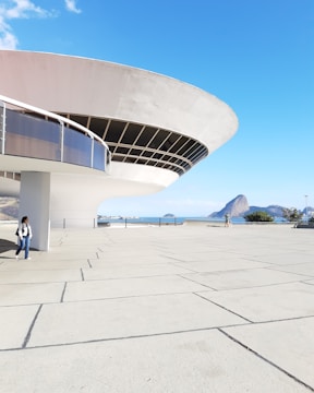A futuristic, disc-shaped building with a sweeping overhang dominates the foreground. The structure features large windows and sleek, modern lines. In the background, a rocky outcrop and a serene blue sea are visible under a clear sky. Two people walk along a spacious, tiled plaza near the building.