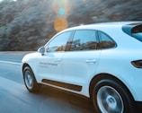 A white SUV is driving quickly on a highway with a blurred background of trees indicating motion. The sunlight creates lens flares that add a dynamic element to the scene. Text in a foreign language is visible on the car's side.