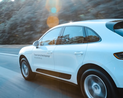 A white SUV is driving quickly on a highway with a blurred background of trees indicating motion. The sunlight creates lens flares that add a dynamic element to the scene. Text in a foreign language is visible on the car's side.