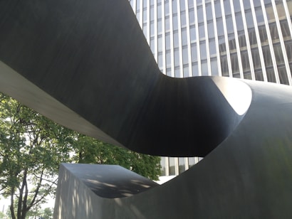 A large abstract metal sculpture with a twisting shape is set in an urban environment. The sculpture contrasts with the stark lines of a modern high-rise building in the background. Leafy trees surround the area, adding a touch of nature to the urban setting.