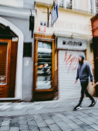 A blurry image of a street scene captures a person wearing a hoodie walking on a cobblestone sidewalk. The background features a storefront with a closed shutter and graffiti. The general atmosphere seems urban and dynamic.