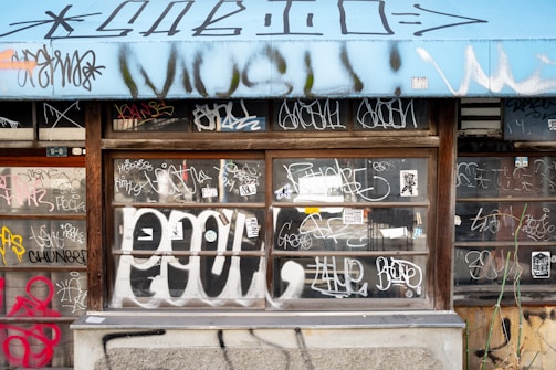 A storefront window extensively covered in various styles of graffiti and tags, set under a blue awning. The window is divided into sections by wooden frames, and the graffiti spans both the glass and surrounding wall areas. The artwork includes a mix of colors and designs, with elements like stickers and abstract shapes.