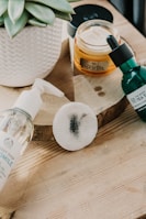 A soft focus image of skincare products arranged on a wooden surface. There is a white potted succulent plant, a jar of face cream labeled 'Oils of Life', a green bottle with a dropper from 'Tea', and a clear pump bottle labeled 'Chamomile'. A round cotton pad with a black smudge is also present.