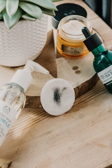 A soft focus image of skincare products arranged on a wooden surface. There is a white potted succulent plant, a jar of face cream labeled 'Oils of Life', a green bottle with a dropper from 'Tea', and a clear pump bottle labeled 'Chamomile'. A round cotton pad with a black smudge is also present.