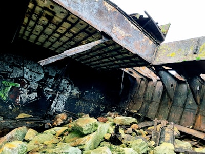 The image features a section of a decaying structure, likely an old shipwreck. Rusted metal beams and panels with visible corrosion and peeling paint create a sense of dereliction. The floor is scattered with large, weathered rocks, and signs of graffiti are visible on the interior walls. The overall environment suggests exposure to harsh elements over time.