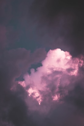 A cloud formation with vibrant pink highlights set against a dark, contrasting sky. The clouds appear soft and fluffy, with the pink tones giving them an ethereal, glowing effect.