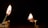 Two matchsticks are burning against a dark background. The left match is positioned on a holder, standing upright with a bright flame. The right match is horizontal, producing a smaller flame and partially supported by a stack of wooden pieces.