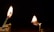 Two matchsticks are burning against a dark background. The left match is positioned on a holder, standing upright with a bright flame. The right match is horizontal, producing a smaller flame and partially supported by a stack of wooden pieces.