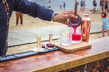 A person is preparing a cocktail at an outdoor bar set up on a wooden table. The drink is vibrant pink and is being poured into a crystal glass through a strainer. Various bar tools and ingredients, including a copper shaker and a small bottle, are arranged on the table. In the background, a sandy beach with several people, including children, is visible.