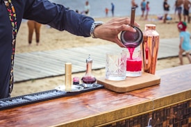 A person is preparing a cocktail at an outdoor bar set up on a wooden table. The drink is vibrant pink and is being poured into a crystal glass through a strainer. Various bar tools and ingredients, including a copper shaker and a small bottle, are arranged on the table. In the background, a sandy beach with several people, including children, is visible.