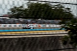 A race car speeds down a track, captured through a chain-link fence. The blurred background suggests high speed, with trees and a large crowd visible in the distance. The car is detailed with designs and sponsorship stickers.