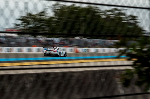A race car speeds down a track, captured through a chain-link fence. The blurred background suggests high speed, with trees and a large crowd visible in the distance. The car is detailed with designs and sponsorship stickers.