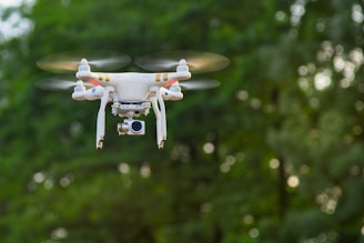 A white quadcopter drone with a camera attached underneath hovers in mid-air against a backdrop of blurred green foliage. The rotors are in motion, suggesting the drone is actively flying.