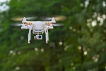 A white quadcopter drone with a camera attached underneath hovers in mid-air against a backdrop of blurred green foliage. The rotors are in motion, suggesting the drone is actively flying.
