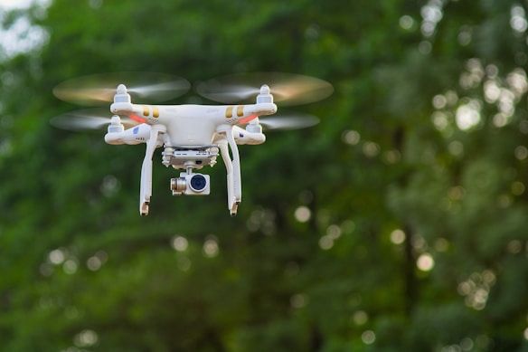 A white quadcopter drone with a camera attached underneath hovers in mid-air against a backdrop of blurred green foliage. The rotors are in motion, suggesting the drone is actively flying.
