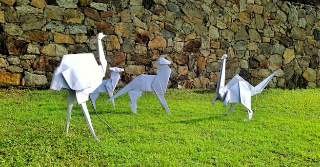Origami sculptures resembling various animals, including a camel and an ostrich, placed on green grass in front of a stone wall.