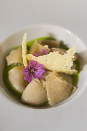 A dish featuring elegantly arranged pasta pieces in a round bowl, accompanied by a vibrant green sauce. The presentation includes a delicate purple flower and a crispy, golden cheese tuile for garnish.