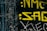 The image features yellow graffiti-style text on a black, textured background. The text appears weathered, suggesting an urban or gritty setting. Scratches and distressed areas are visible, adding a rough aesthetic.