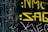 The image features yellow graffiti-style text on a black, textured background. The text appears weathered, suggesting an urban or gritty setting. Scratches and distressed areas are visible, adding a rough aesthetic.