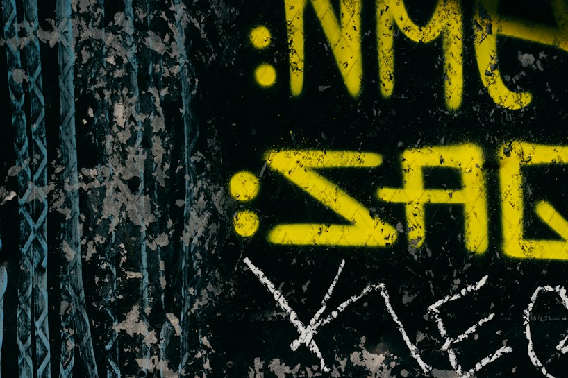The image features yellow graffiti-style text on a black, textured background. The text appears weathered, suggesting an urban or gritty setting. Scratches and distressed areas are visible, adding a rough aesthetic.