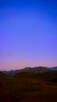 A serene landscape featuring rugged mountains with a clear twilight sky transitioning from deep blue to soft pink. The foreground consists of dark hills, and small white structures are visible in the distance, possibly tents or caravans.
