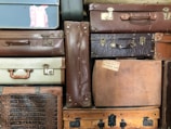 A stack of vintage suitcases in various materials and colors, including leather, fabric, and textured surfaces. There are metal clasps, handles, and labels on the suitcases, hinting at their use in travel. A visible luggage label indicates a journey from Hayling Island to Bournemouth Central.