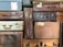 A stack of vintage suitcases in various materials and colors, including leather, fabric, and textured surfaces. There are metal clasps, handles, and labels on the suitcases, hinting at their use in travel. A visible luggage label indicates a journey from Hayling Island to Bournemouth Central.