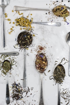 Various types of loose tea leaves and herbs are displayed on metal spoons against a white marble background. The spoons are scattered across the surface, each holding different tea blends, including chamomile flowers, green tea, and other herbal mixes.