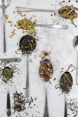Various types of loose tea leaves and herbs are displayed on metal spoons against a white marble background. The spoons are scattered across the surface, each holding different tea blends, including chamomile flowers, green tea, and other herbal mixes.