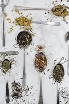 Various types of loose tea leaves and herbs are displayed on metal spoons against a white marble background. The spoons are scattered across the surface, each holding different tea blends, including chamomile flowers, green tea, and other herbal mixes.