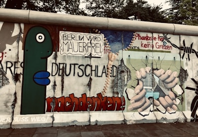 Artwork on a section of the Berlin Wall featuring bold graffiti with German text, a large green cartoon figure, and a depiction of hands breaking through the wall. The scene includes barbed wire and imagery suggesting freedom and liberation.