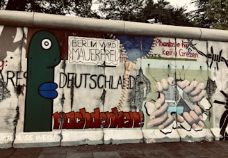 Artwork on a section of the Berlin Wall featuring bold graffiti with German text, a large green cartoon figure, and a depiction of hands breaking through the wall. The scene includes barbed wire and imagery suggesting freedom and liberation.