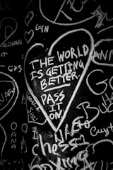 Chalk writing on a black surface features a heart-shaped outline containing the message 'The world is getting better. Pass it on.' Surrounding the central message are various random words and scribbles in different orientations.