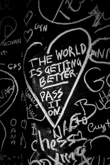 Chalk writing on a black surface features a heart-shaped outline containing the message 'The world is getting better. Pass it on.' Surrounding the central message are various random words and scribbles in different orientations.