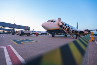 An airplane is parked on the tarmac with passengers boarding via a staircase. The airport signage is visible in the background, and there is a service vehicle nearby. The sky is clear with a hint of the evening or morning light.