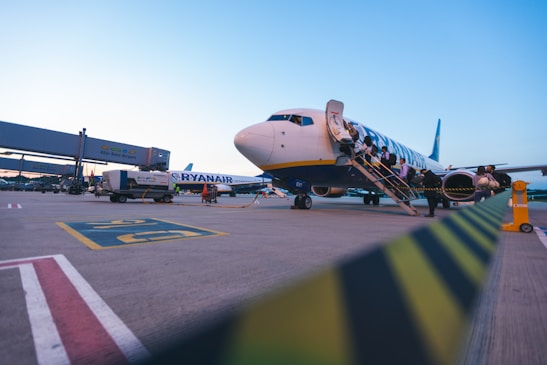 An airplane is parked on the tarmac with passengers boarding via a staircase. The airport signage is visible in the background, and there is a service vehicle nearby. The sky is clear with a hint of the evening or morning light.