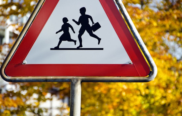 A triangular road sign with a red border featuring a silhouette depiction of two children running, set against a blurred background of autumn leaves.