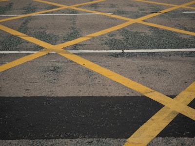 An asphalt road surface features a grid of yellow crosshatched lines, with white lines intersecting horizontally. The surface is weathered, showing signs of wear and patches of darker textures.