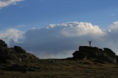A person stands triumphantly on a large rock formation under a vast open sky filled with cumulus clouds. The silhouette of the individual with raised arms creates a sense of achievement against the expansive backdrop.