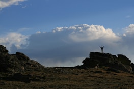 A person stands triumphantly on a large rock formation under a vast open sky filled with cumulus clouds. The silhouette of the individual with raised arms creates a sense of achievement against the expansive backdrop.