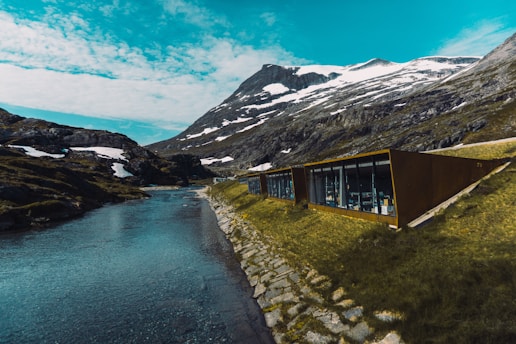 A modern glass building nestled in a mountainous landscape, positioned beside a narrow river. Snow patches are visible on the rugged mountain slopes under a partly cloudy sky.