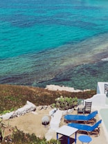 The scene features a picturesque coastal setting with vibrant turquoise waters meeting the land. A patio area with blue lounge chairs, a small table, and a couple of black chairs is visible in the foreground. The area is bordered by white walls and greenery, including cactus plants. Rocks and the clear sea are prominent features, creating a serene, inviting atmosphere.